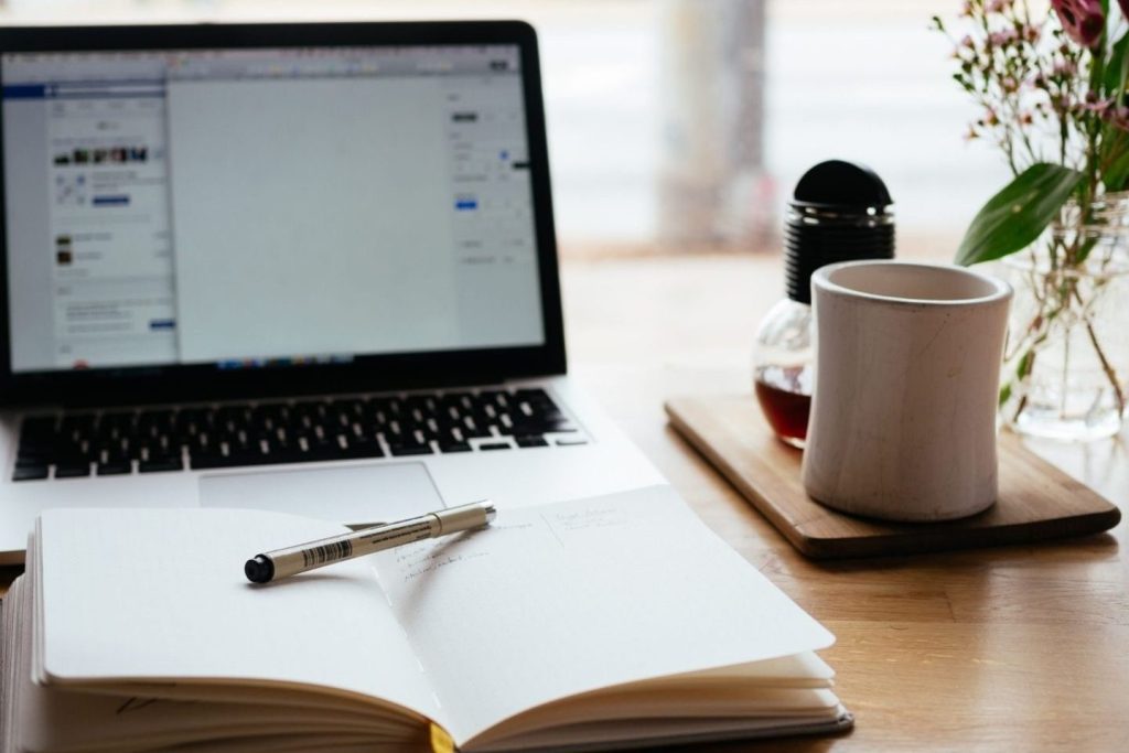 Open notebook with a pen placed on top in front of a laptop on a wooden desk.