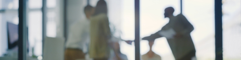 Blurred silhouette-style view of multiple people meeting in a glass-walled office, with one person gesturing during a discussion.
