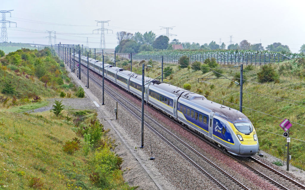 Eurostar high-speed train travelling along electrified railway tracks through a rural landscape, viewed from an elevated position. Power lines, vegetation, and open countryside surround the rail corridor.